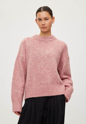 Jeune femme aux cheveux foncés attachés en chignon, portant un pull en maille rose ample et un pantalon noir plissé sur un fond clair uni.