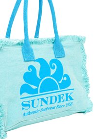 Bolsa de tela azul con bordes con flecos, que presenta un logo de sol y ola en azul, etiquetada "SUNDEK Ropa de Surf Auténtica Desde 1958."