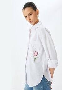 Camicia bianca con bottoni, dotata di tasca con un design di tulipano rosa e piccoli elementi decorativi. Tessuto morbido con un motivo texturizzato.