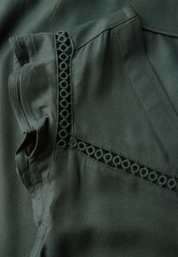 Ciemnozielony materiał o miękkiej teksturze z dekoracyjnym wykończeniem w postaci okrągłych wycięć przy dekolcie, z gładkimi brzegami i szwami.