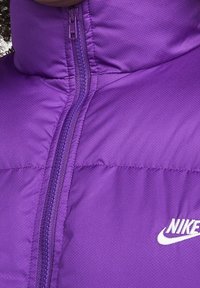 Fialová zateplená bunda s předním zipem a bílým logem Nike na hrudi, kterou nosí osoba s kudrnatými vlasy.