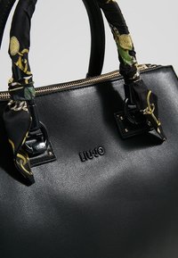 Sac à main en cuir noir texturé avec fermeture éclair dorée, foulards décoratifs noirs et dorés à motifs enroulés autour des poignées arrondies, logo LIU JO sur le devant.