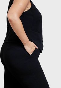 Svart ärmlös topp med sidofickor, skräddarsydd passform, med en slät textur och strukturerade sömmar, visad från sidan.