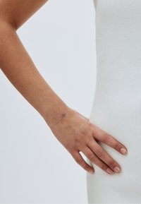 Sidabrinė grandinėlė su maža kryžiaus pakabuku, pasižyminti subtiliomis jungtimis, minimalistinio dizaino, dėvima ant rankos prieš šviesų foną.