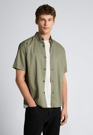 Junger Mann trägt ein aufgeknöpftes, olivgrünes Hemd mit kurzen Ärmeln über einem weißen T-Shirt und schwarzen Hosen, steht vor einem schlichten hellen Hintergrund.