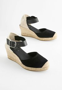 Scarpe nere in suede con zeppa, schiena intrecciata beige, cinturino elastico alla caviglia e punta beige. La suola intermedia in iuta stile espadrille offre texture.