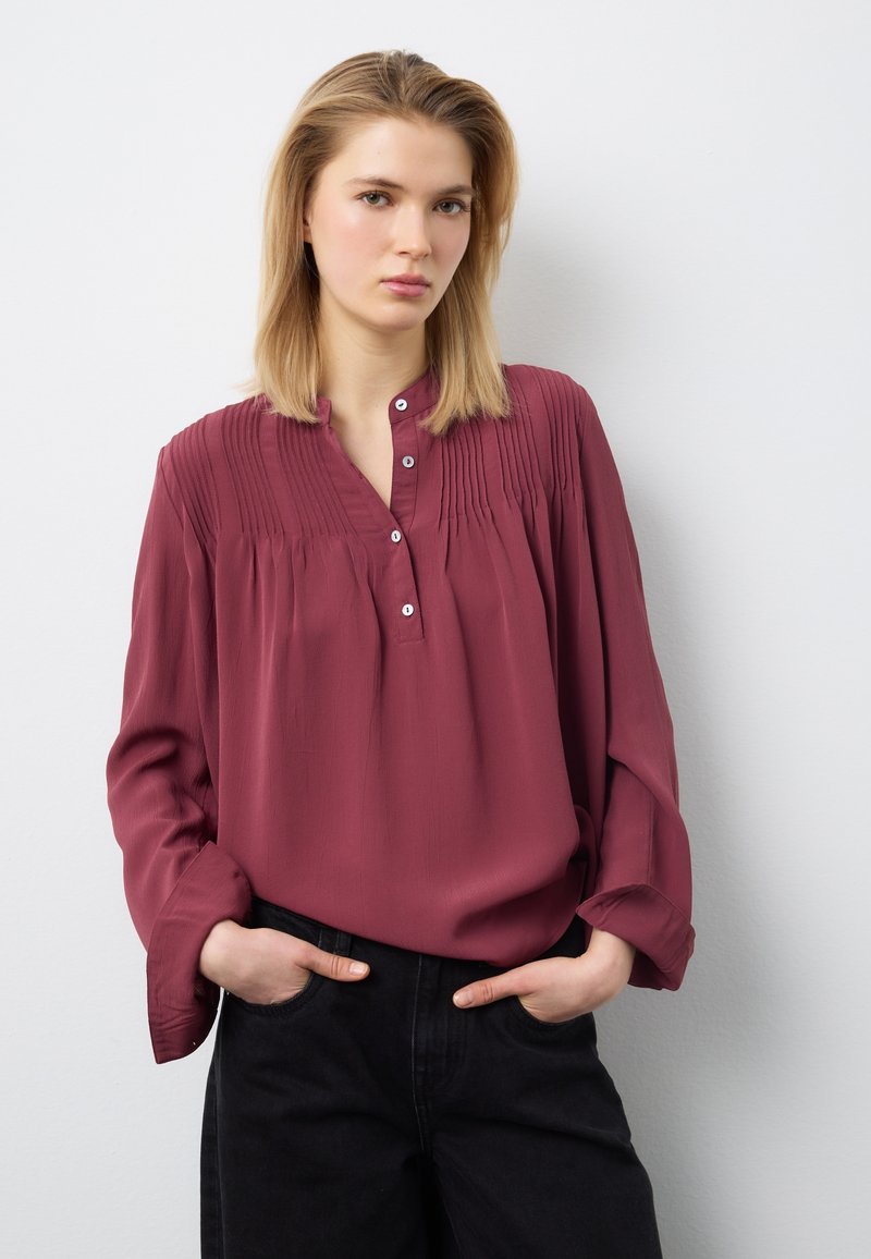 Junge Frau mit glattem blondem Haar, die eine bordeauxrote Bluse mit langen Ärmeln und schwarze Hose trägt, die Hände in den Taschen, vor weißem Hintergrund stehend.