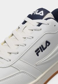 Baskets en cuir blanc avec logo FILA bleu marine et lacets assortis, présentant une partie supérieure texturée et une semelle extérieure partiellement en gomme sur fond blanc.