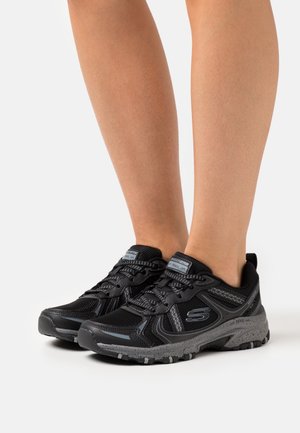 Jambes portant des chaussures de course sur sentier Skechers noires et grises avec semelles texturées sur fond blanc.