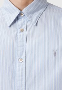 Lysblå stribet skjorte lavet af glat stof. Har en button-down krave og to knaplukninger, med et diskret broderet gedelogo.
