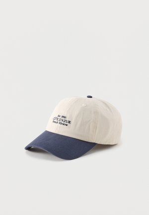 Baseball cap with cream crown and navy blue brim, embroidered text "Est. 1950 Côte d'Azur French Riviera" on front panel.
