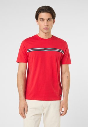 Giovane uomo con capelli corti e scuri che indossa una maglietta rossa con strisce orizzontali nere, bianche e rosse sul petto e pantaloni beige chiaro.