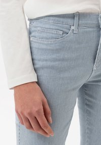 Svetle modré pruhované džíny s úzkým střihem, které mají pět kapes, zapínání na knoflík a jemné detaily šití. Hladká textura.