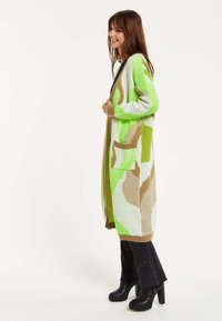 Long cardigan in vibrant green, beige, and cream. Knit texture with open front, two pockets, and ribbed cuffs. Paired with black flared jeans.