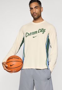 Dlhý rukáv krémového športového trička s nápisom „Cream City“ v zelenej farbe, modré a zelené bočné pruhy, v kombinácii so sivými šortkami, držiaci basketbalovú loptu.