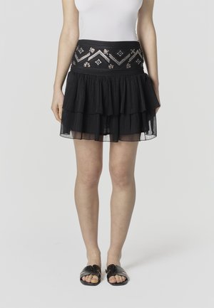 Mujer con falda negra de varias capas con un patrón de lentejuelas en la cintura, camiseta blanca y sandalias negras con tachuelas y punta abierta, de pie sobre un suelo blanco.