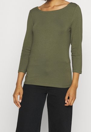 Femme portant un haut vert olive à manches longues et col bateau, ainsi qu'un pantalon noir, debout avec les bras détendus sur un fond neutre.