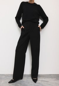 Pull noir à manches longues associé à un pantalon noir taille haute et des escarpins noirs à bout pointu. Textures lisses et design minimaliste.