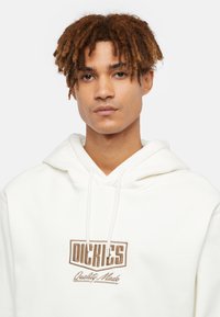 Witte hoodie van zacht materiaal met een voorzak. Bevat een bruine geborduurde logo "DICKIES" met daaronder "Quality Made".