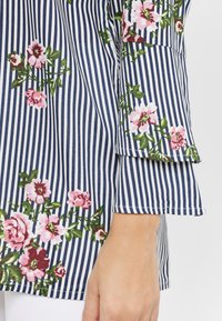 Navy- och vitrandig tyg med mönster av rosa och gröna blommor. Har en utvändig ärm och en slät textur.
