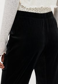 Zwarte fluwelen broek met een brede, elastische tailleband. De stof heeft een gladde, glanzende textuur. Een witte, geribbelde, lange mouwtop is gedeeltelijk zichtbaar.
