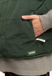 Grön quiltad väst med en dragkedjeficka där en etikett syns, tillverkad av ett lättviktigt material. Lagd över en grå sweatshirt.