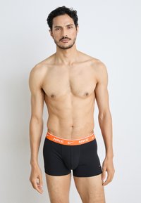 Schwarze Boxershorts mit einem orangefarbenen Nike-Logo-Bund, die eine enge Passform und weiches Baumwollmaterial aufweisen, präsentiert von einem Mann mit muskulösem Körperbau.
