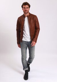 Giacca in pelle marrone con una finitura leggermente texture, chiusura con zip e tasche laterali, indossata sopra una camicia bianca e abbinata a jeans azzurri chiari.