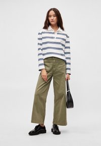 Ριγέ πουκάμισο με μακρύ μανίκι σε λευκό και ναυτικό μπλε, παντελόνι φαρδύ σε ελιά, και μαύρα γυαλιστερά παπούτσια, κρατώντας μια μικρή μαύρη τσάντα.