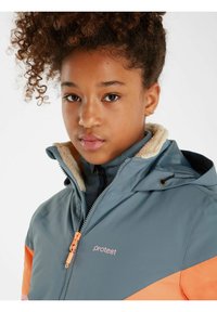Veste imperméable avec un corps teal, des accents orange et une doublure en polaire. Dispose d'une fermeture éclair, d'une capuche ajustable et d'un logo sur la poitrine.