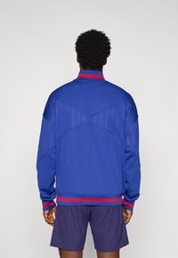 Mand iført en blå jakke med røde striber på kraven og ærmerne samt mørkeblå shorts, vist fra baggrunden mod en ensfarvet baggrund.