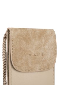 Beige läder crossbody-väska med en texturerad mockaflap och präglad "EXPATRIÉ"-logotyp. Har en flätad rem och en slät yta.