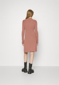 Robe côtelée rose, longueur genou, manches longues, avec des accents dorés aux épaules et un col rond. Portée avec des bottines noires.