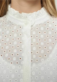 Blusa blanca bordada con un cuello con volantes, cierre de botones y un patrón floral con recortes de ojal en toda la tela.