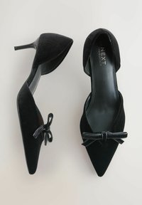 Sorte spidse højhælede sko med fløjlstekstur og små dekorative sløjfer, vist fra top- og sidevinkel på en lys baggrund.