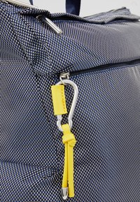 Bolsa de tela texturizada en azul marino con un acento de cuero amarillo y un mosquetón plateado. Presenta un cierre de cremallera y un diseño estructurado.