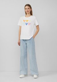 T-shirt en coton blanc avec des graphismes de coeurs colorés en orange, jaune, bleu et rose, associé à un jean ample bleu clair et des baskets blanches.