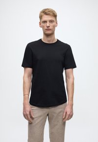 Schwarzes Baumwoll-T-Shirt mit kurzen Ärmeln, Rundhalsausschnitt und geradem Saum. Das Modell kombiniert es mit hellbeigen, strukturierten Hosen.