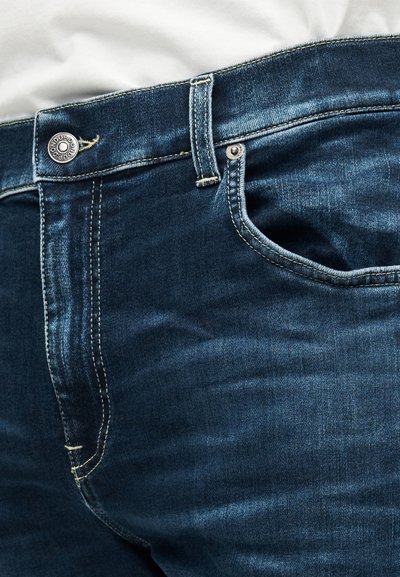 Nærbillede af mørkeblå denimjeans med linning, knap og forlomme med synlige sømme og metalnitter mod en hvid stofbaggrund.