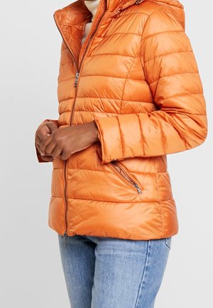 Personne portant une veste matelassée orange fermée, les mains jointes à la taille et un jean bleu clair visible.