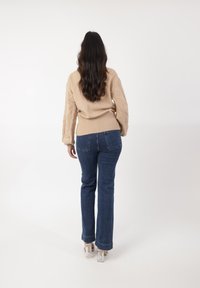 Beżowy sweter dziany z teksturowymi rękawami, w połączeniu z niebieskimi dżinsami w stylu dzwonów i przezroczystymi butami na obcasie, widok z tyłu.