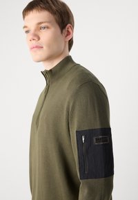 Olivgrüner Zip-Kragen-Pullover aus weichem Stoff, mit einer schwarzen Reißverschluss-Tasche am linken Ärmel und einem strukturierten Patch mit Markenlogo.
