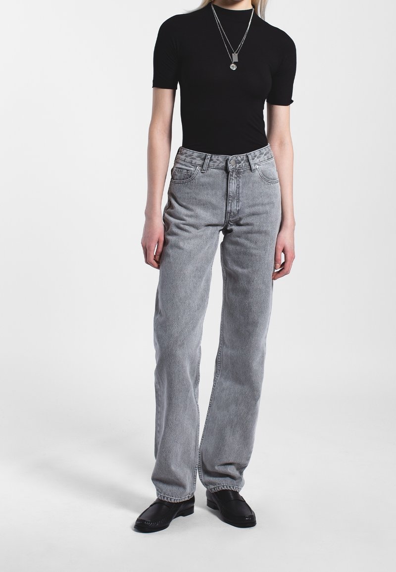 Personne portant un haut noir à manches courtes, des colliers argentés superposés, un jean gris taille haute à jambes droites, et des chaussures noires sans lacets sur fond blanc.