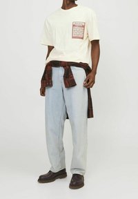 Man som bär en beige t-shirt med en grafisk applikation, ljusblå jeans, mörkbruna stövlar och en röd och svart rutig skjorta knuten runt midjan.