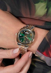 Reloj de pulsera en tonos dorado y plateado con una esfera verde, números romanos, ventana de fecha y acentos de diamantes en una correa de metal trenzado.