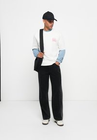 T-shirt blanc superposé à une chemise à manches longues bleue, pantalon baggy noir et casquette noire. Sac noir porté en bandoulière. Baskets beiges.