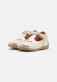 Pequeños zapatos Mary Jane metálicos dorados para niños con diseños perforados en la parte frontal y correas de Velcro sobre un fondo liso.
