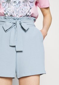 Shorts bleu clair avec une taille froncée et un accent en nœud noué. Comprend des poches latérales et un tissu lisse et texturé.