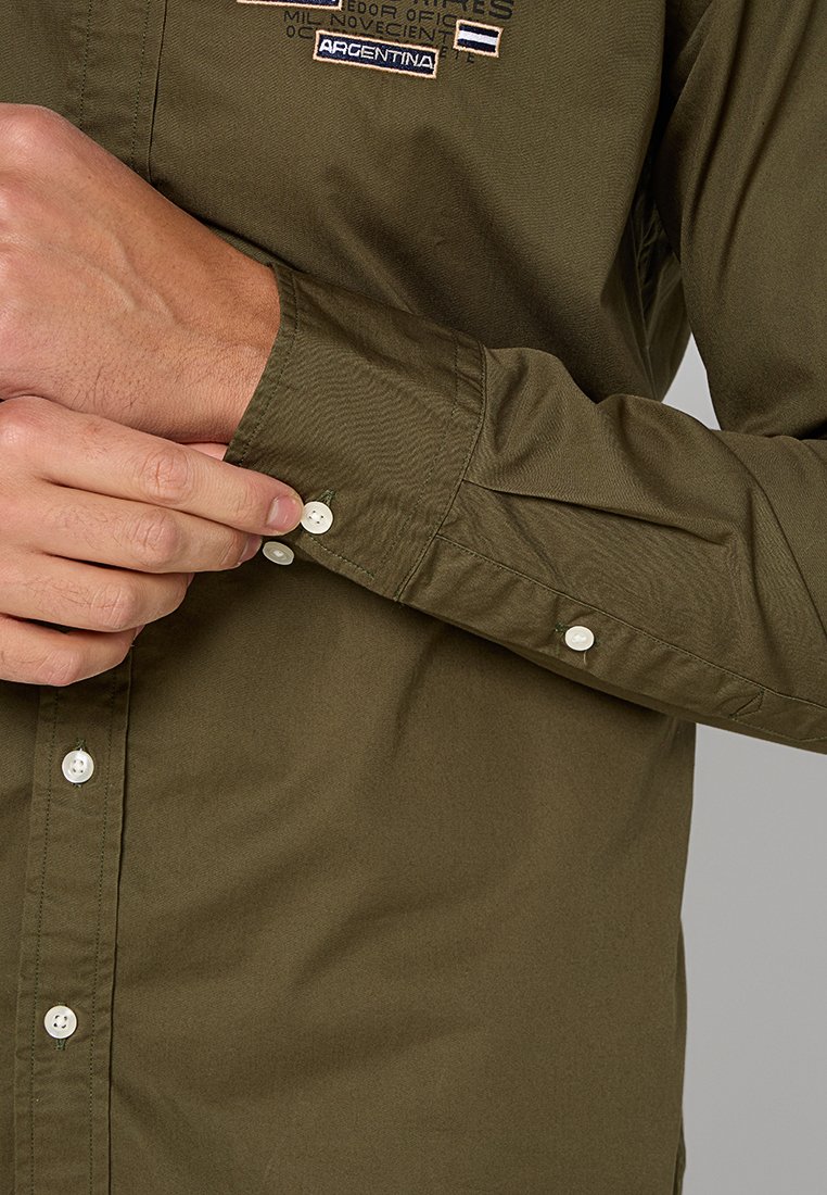 Persona che allaccia un bottone su una camicia a maniche lunghe verde oliva con una patch ricamata "Argentina" sul petto.