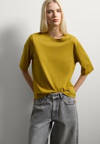 T-shirt a maniche corte color giallo senape realizzato in materiale morbido, abbinato a jeans in denim grigio a vita alta con una vestibilità rilassata e tasche.